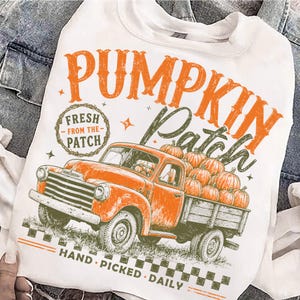 Op de afbeelding: Een wit t-shirt met een afbeelding van een oranje vintage pick-up truck geladen met pompoenen. De tekst luidt PUMPKIN PATCH, FRESH FROM THE PATCH en HAND PICKED DAILY. Het ontwerp bevat oranje en olijfgroene kleuren met een geblokte rand.