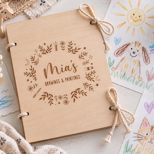 Op de afbeelding: Een rechthoekig schetsboek van lichtgekleurd hout met een touwsluiting. De kaft is voorzien van een lasergegraveerd bloemenkransontwerp rondom de tekst "Mia's DRAWINGS & PAINTINGS" in een donkerbruin lettertype.