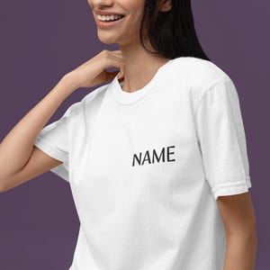 May include: White t-shirt with the word "NAME" printed in black. The shirt has a crew neck and short sleeves. The model is smiling and wearing the shirt.