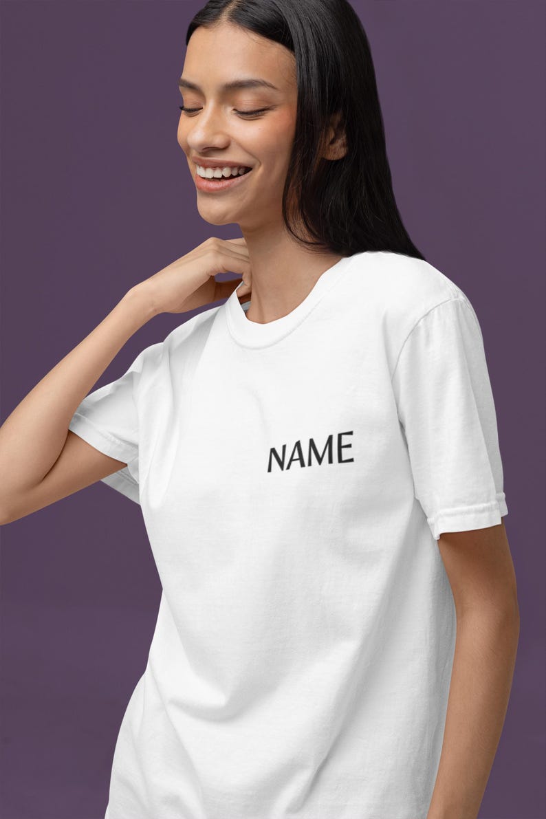 May include: White t-shirt with the word "NAME" printed in black. The shirt has a crew neck and short sleeves. The model is smiling and wearing the shirt.