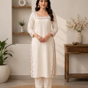 Op de afbeelding: Een crèmekleurige lange kurta met driekwartmouwen en een bijpassende broek met rechte pijpen. Het kledingstuk heeft een vierkante halslijn en zijsplitten, beide versierd met delicaat bloemenborduursel in roze, blauwe en groene tinten.