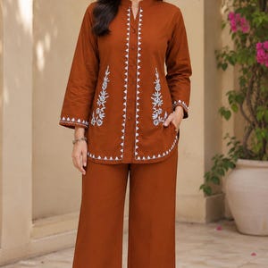 May include: A rust-colored two-piece set consisting of a long-sleeved tunic and wide-leg trousers. The tunic features a mandarin collar, a central button placket, and white floral embroidery along the front, hem, and cuffs. The trousers are solid.