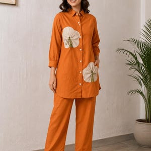 May include: A burnt orange long-sleeve button-down shirt featuring two large, beige, circular fabric appliqués with small green tassels, paired with matching burnt orange wide-leg trousers. The shirt has a classic collar and white buttons.