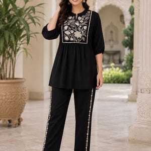 May include: A black tunic and matching trousers set. The tunic features three-quarter sleeves, a split neckline, and a cream-coloured floral embroidery panel on the chest. The trousers include a vertical cream-coloured floral embroidery stripe on each leg.
