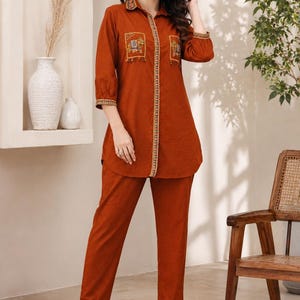 Op de afbeelding: Een roestkleurige tweedelige set bestaande uit een tuniek met knoopsluiting en een bijpassende broek. De tuniek heeft een kraag, driekwartmouwen en twee borstzakken met geborduurde olifantenmotieven en een decoratieve bies langs de sluiting.