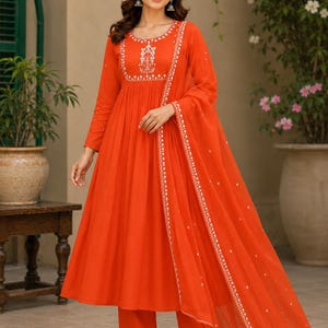 Può includere: Un abito arancione vivace in stile Anarkali con maniche lunghe, scollo rotondo con ricami floreali bianchi e vita arricciata. L'outfit include pantaloni dritti abbinati e una dupatta arancione trasparente con bordi ricamati in bianco.
