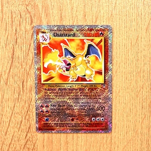 Może przedstawiać: Holograficzna karta kolekcjonerska Charizard przedstawiająca pomarańczowego, smoczego stwora z niebieskimi skrzydłami ziejącego ogniem. Karta zawiera nazwę Charizard, 120 HP, moc Energy Burn oraz atak Fire Spin o sile 100 obrażeń, wydrukowane czarną czcionką.