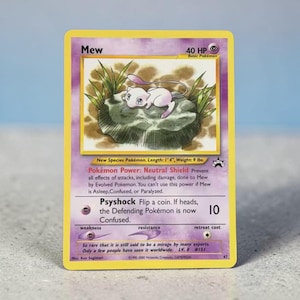 May include: A Mew trading card with a yellow border and purple background. The illustration shows a small pink creature resting on a green rock. Text includes Mew, 40 HP, Neutral Shield, Psyshock, and details about its length of 1'4" and weight of 9 lbs.