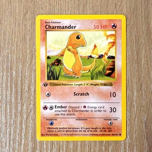 May include: A Charmander Pokémon trading card with a yellow border. The card features an orange Charmander character with a flame in the background. The card text includes "Scratch" and "Ember" attack details, and the text "Lizard Pokémon. Length: 2' 0", Weight: 19 lbs."