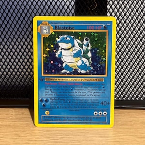 May include: A holographic Blastoise trading card with a yellow border. The card displays the character Blastoise, 100 HP, and the text Shellfish Pokémon, Length: 5' 3", Weight: 189 lbs. It includes the moves Rain Dance and Hydro Pump.
