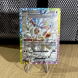 May include: A holographic Eevee ex trading card featuring a Tera crystal design with a rainbow border. The card displays the character Eevee, the Rainbow DNA ability, and the Coruscating Quartz attack with 200 HP, set against a shimmering background.