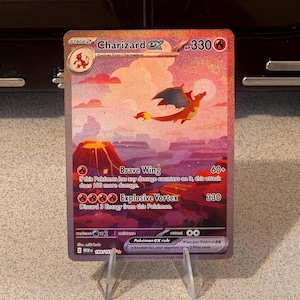 May include: A Charizard ex trading card featuring a vibrant illustration of the fire-type Pokémon flying over a volcanic landscape. The card displays 330 HP, the Brave Wing and Explosive Vortex attacks, and the set number 199/165.