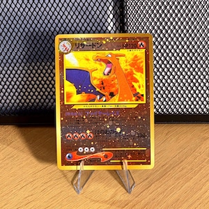 May include: A gold-colored Charizard trading card featuring a fiery orange dragon illustration, HP120 text, and Japanese characters. The card displays four fire energy symbols, a retreat cost of three colorless symbols, and a dark background.