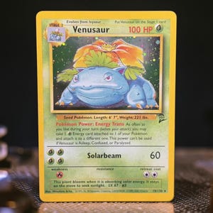 May include: A holographic Venusaur trading card with a yellow border. The card displays the character, 100 HP, the Energy Trans Pokémon Power, and the Solarbeam attack. Text includes Seed Pokémon, Length 6' 7", Weight 221 lbs, and LV. 67 #3.