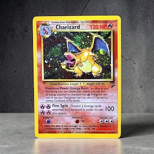 May include: A Charizard trading card featuring a holographic illustration of the orange dragon-like creature with blue wings. The card displays 120 HP, the Energy Burn Pokémon Power, and the Fire Spin attack. Text includes length 5' 7" and weight 200 lbs.