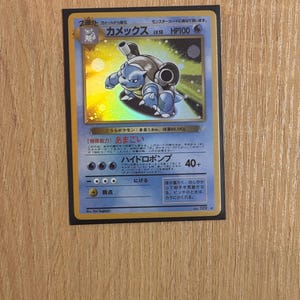 Puede incluir: Una carta coleccionable Pokémon holográfica con Blastoise, una criatura azul con forma de tortuga y cañones. La carta tiene texto japonés y 100 PS. La carta está en una funda negra.