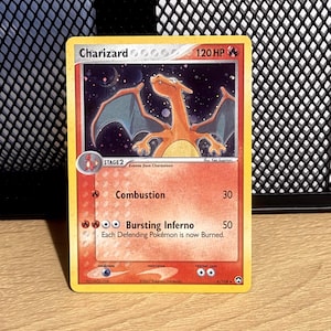 May include: A Charizard trading card with 120 HP, featuring an orange dragon-like creature against a starry night sky. The card displays the attacks Combustion and Bursting Inferno, with text detailing the effects of the moves and stage information.