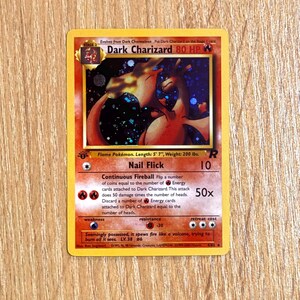 May include: A Dark Charizard Pokémon card with a fiery illustration. The card features the text "Dark Charizard 80 HP" and "Nail Flick 10". The card's dimensions are 170 cm in length and weighs 90 kg. The card is a stage 2 card.