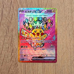 Op de afbeelding: Een holografische Pikachu EX Pokémon-verzamelkaart. De kaart toont een gele Pikachu-personage met een kleurrijk geometrisch ontwerp en Japanse tekst. De kaart heeft een roze en gouden achtergrond.