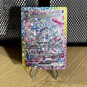 May include: A holographic Sylveon ex trading card featuring a pink and blue character illustration, 270 HP, and the attacks Magical Charm and Angelite. The card has a rainbow-colored, shattered-glass foil finish and displays text detailing its abilities and game rules.