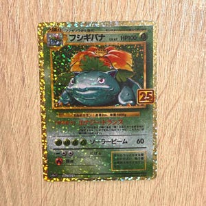 Könnte beinhalten: Holografische Pokémon-Sammelkarte mit einem grünen Bisaflor und einer rot-orangen Blume auf dem Rücken. Die Karte hat japanischen Text, ein "25"-Logo und eine HP100-Bewertung. Die Karte liegt auf einer Holzoberfläche.