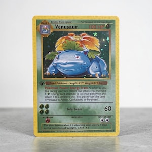 May include: A holographic Venusaur trading card with a yellow border, featuring the blue creature with a large flower on its back. The card displays 100 HP, the Energy Trans Pokémon Power, the Solarbeam attack, and text including length 6' 7" and weight 221 lbs.