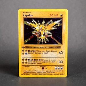May include: A yellow Zapdos trading card featuring a holographic illustration of the electric bird Pokémon. The card displays 90 HP, the attacks Thunder and Thunderbolt, and text detailing its length of 5' 3" and weight of 116 lbs.