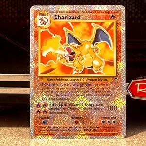May include: A holographic Charizard trading card featuring the Stage 2 Flame Pokémon with 120 HP. The card displays the Energy Burn Pokémon Power and Fire Spin attack, with text detailing a length of 5' 7" and weight of 200 lbs.