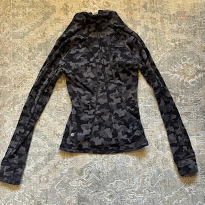 Op de afbeelding: Een sportshirt met lange mouwen en een donkergrijs en zwart camouflagepatroon. Het kledingstuk heeft een hoge kraag, een getailleerd silhouet met verticale naaddetails en mouwboorden met duimgaten. Een klein rond logo is linksonder zichtbaar.