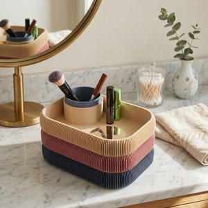 May include: A stack of three ribbed trays in beige, red, and navy blue, holding makeup brushes and containers. A small mirror and beauty products are also visible. The trays are on a marble surface, with a mirror and decorative items in the background.