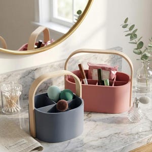 May include: Two cosmetic organizers with wooden handles. One is dusty rose, the other is blue-gray. Each has internal dividers. The blue-gray organizer holds makeup sponges and a brush. The dusty rose organizer holds makeup items. A marble countertop is in the background.