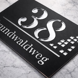 May include: A black rectangular address sign with the number "38" and the word "Grundwaldweg" in a silver, cut-out design. The sign has a modern, minimalist aesthetic.