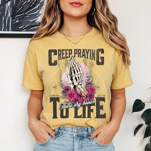 May include: A mustard yellow short-sleeve t-shirt featuring a graphic of skeletal hands in a prayer position surrounded by pink and purple flowers. The text reads CREEP PRAYING TO LIFE and EZEKIEL 37:1-10 Those dry bones.