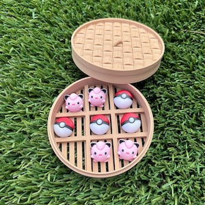 May include: A tan, circular, 3D-printed bamboo steamer container featuring a tic-tac-toe grid insert. The game set includes four pink Jigglypuff character figurines and five red and white Poké Ball game pieces arranged within the grid.
