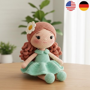 Könnte beinhalten: Eine handgefertigte Häkelpuppe mit einem hellrosa Gesicht, braunen Locken und einem weißen Gänseblümchen-Haarschmuck. Die Puppe trägt ein mintgrünes Kleid und hat passende runde Füße. Die Puppe sitzt auf einer Holzoberfläche.