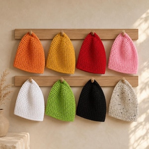 May include: Eight crochet beanies in orange, yellow, red, pink, white, green, black, and speckled beige, each hanging on a wooden peg rack. The hats feature a consistent textured stitch pattern and are arranged in two rows of four on the wall.