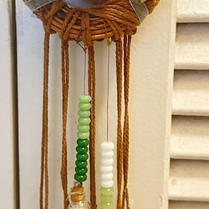 Peut inclure: Un heurtoir de porte décoratif avec une base en couronne tressée, orné de roses beiges et de ruban argenté. Des cordons bruns pendent, suspendant des brins de perles vertes et blanches, de petites cloches et des pendentifs en forme de jade.