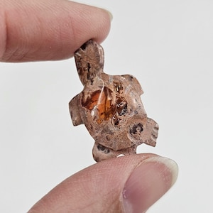 May include: A small, carved turtle figurine held between fingers. The turtle is a mix of brown and orange hues, with a textured surface. The figurine's design is detailed, showcasing the turtle's shell and head.
