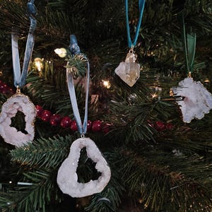 May include: Four crystal geode ornaments with gold trim hang from blue and green ribbons on a Christmas tree. The ornaments have a natural, irregular shape and are various shades of white, gray, and clear.