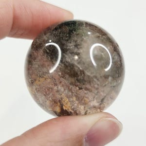 May include: A clear, round crystal ball with internal inclusions of brown, green, and reddish hues. The polished sphere is held between fingers, showcasing its translucent quality and natural patterns. The crystal ball is a decorative item.