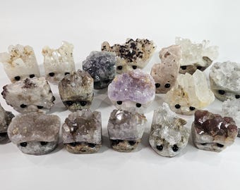 May include: A collection of small, carved crystal figurines with black eyes. The crystals vary in color, including clear, purple, and brown. The figurines are arranged on a white surface.