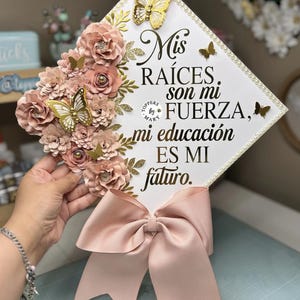 May include: A graduation cap with a white surface, adorned with paper flowers, gold butterflies, and the text "Mis raíces son mi fuerza, mi educación es mi futuro." A large pink bow is attached to the cap.