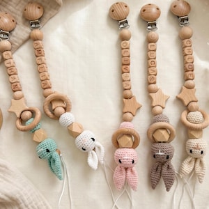 Könnte beinhalten: Baby-Schnullerclips aus Holz mit Namen, die in Holzperlen geschrieben sind. Jeder Clip hat ein anderes gehäkeltes Tier, darunter ein Hase und ein Oktopus. Die Clips sind in verschiedenen Farben erhältlich, darunter Rosa, Grün und Beige.