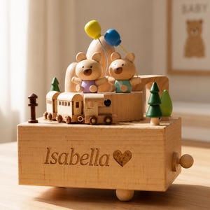 May include: A wooden music box featuring two small bears, a train, and two balloons in yellow and blue. The front of the box is engraved with the name Isabella and a heart symbol. The design includes miniature green trees and a brown post.