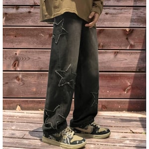 May include: Black denim jeans with large, distressed star patches. The jeans have a relaxed fit and are paired with camouflage sneakers. The background is a wooden wall.