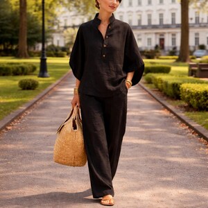 Puede incluir: Un conjunto de lino negro, que incluye una camisa con botones y pantalones de pernera ancha, se combina con un bolso tote tejido. La camisa tiene cuello mandar&iacute;n y tres botones. La modelo lleva pulseras doradas y sandalias, caminando por un camino pavimentado.