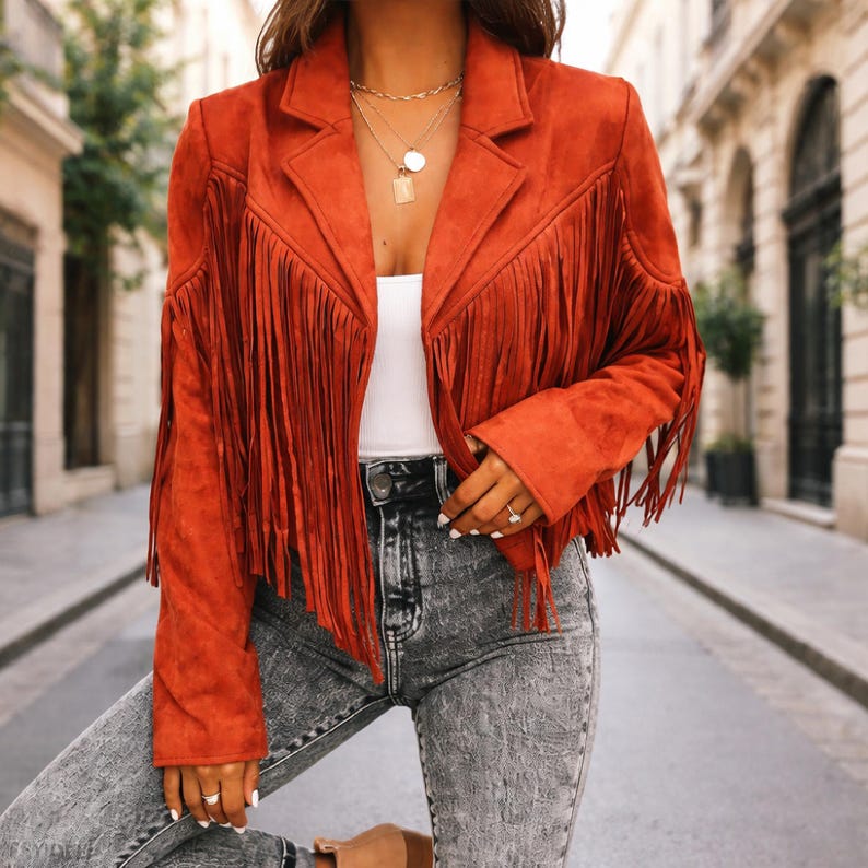 May include: Rust-colored suede jacket with fringe detailing. The jacket features a classic collar and long sleeves, with fringe accents along the front and sleeves. The jacket is paired with gray denim jeans.