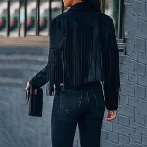 May include: Black suede jacket with fringe detail on the back and sleeves. The jacket has a classic collar and a cropped fit. The model is wearing black jeans and holding a black clutch.