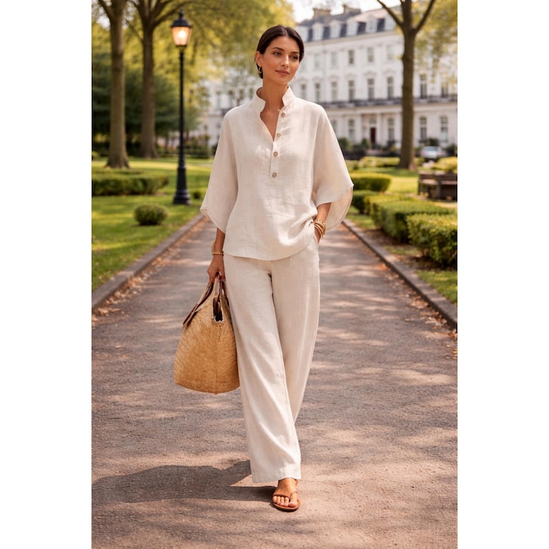 Puede incluir: Una mujer con un conjunto de lino color crema, que incluye una camisa con botones y pantalones de pierna ancha. Lleva un bolso tote tejido y sandalias. El atuendo es de tonos neutros, adecuado para el clima c&aacute;lido. El fondo muestra un parque.