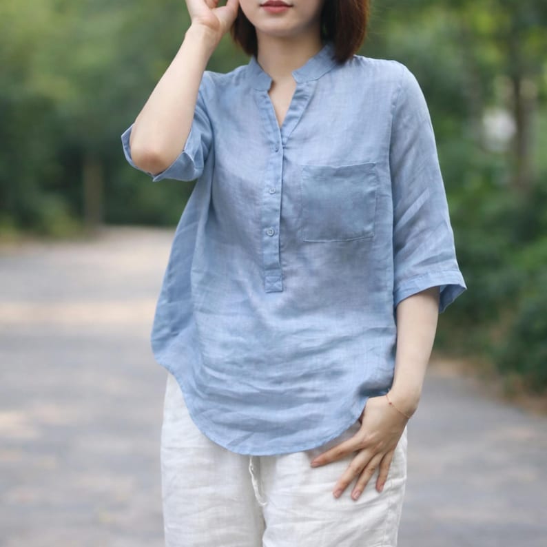 May include: A pale blue linen shirt with a mandarin collar and a single chest pocket. The shirt has short sleeves and a relaxed fit. The model is wearing white linen trousers. The background is a blurred outdoor scene.